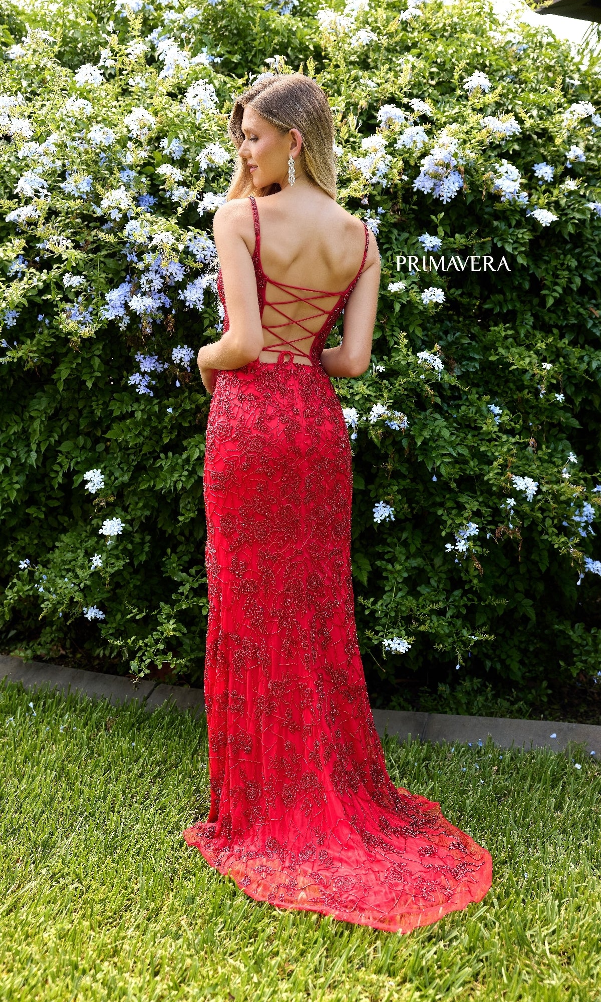A woman wearing the Primavera 4317 Lace-Up Long Lace Prom Dress in red stands on grass before a bush with white flowers.